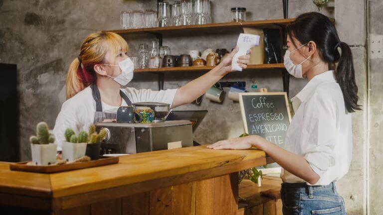 young-female-restaurant-staff-wearing-protective-face-mask-using-infrared-thermometer-checker-or-temperature-gun-on-customer-s-forehead-before-enter-768x432(1)